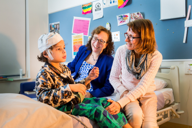 A child wearing a hospital gown and a cap sits on a hospital bed, talking with 2 smiling adults beside them in a room with colourful decorations and posters on the wall.