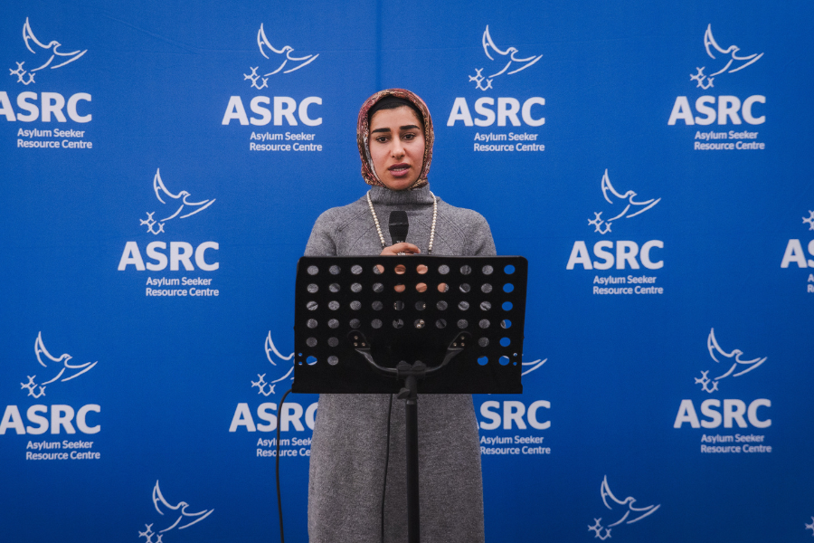 Asylum seeker standing at a podium with a microphone, speaking at the Asylum Seeker Resource Centre, with a backdrop featuring the centre's logo.