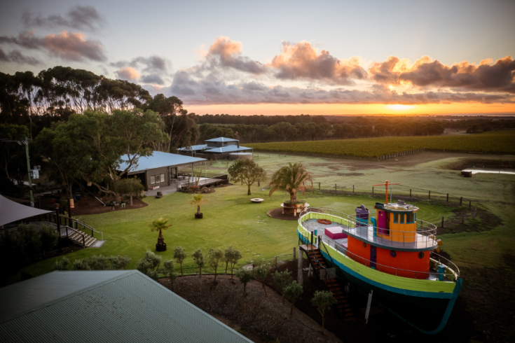 Sunset view over landscape featuring a playground with a colourful boat structure, green lawns, and distant vineyards.