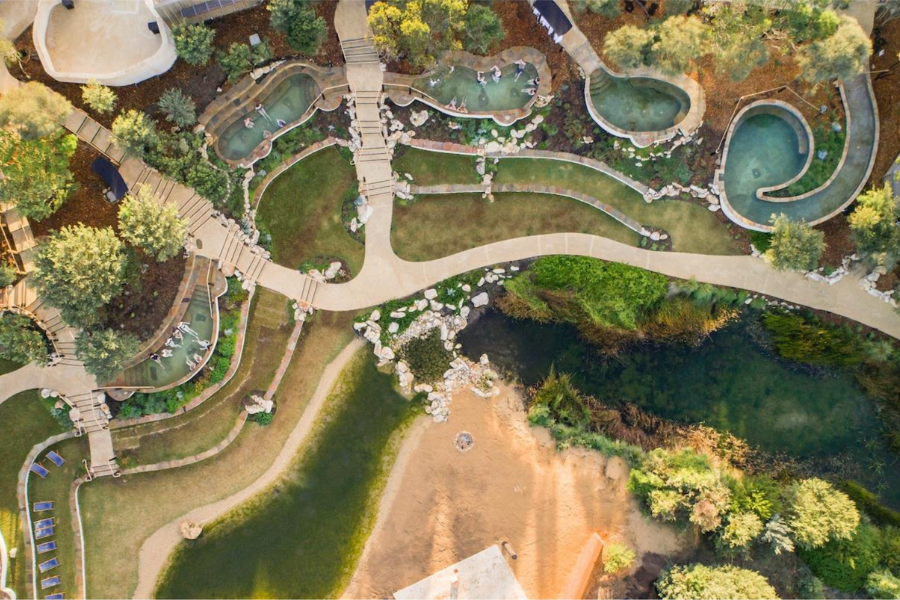 Peninsula Hot Springs This is an aerial view of a lush, landscaped area featuring winding pathways, small pools, and rock formations surrounded by greenery. The scene includes a mix of pathways, vegetation, and hot springs.