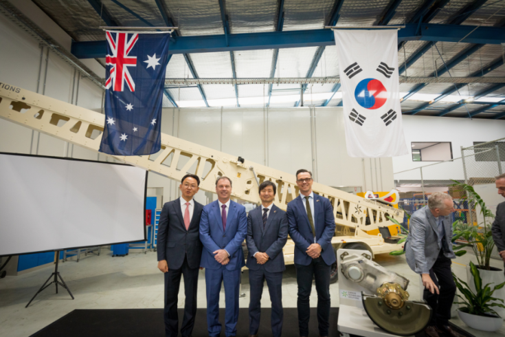 Representatives from Corvus Technology Solutions and the Victorian Government standing formally in a warehouse with the Australia and South Koren flags hanging from the roof.