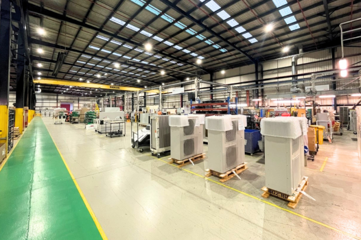 A spacious industrial facility with high ceilings and bright overhead lights. The area contains multiple large air conditioning units placed uniformly on wooden palates. The overall environment is clean and well-organised.