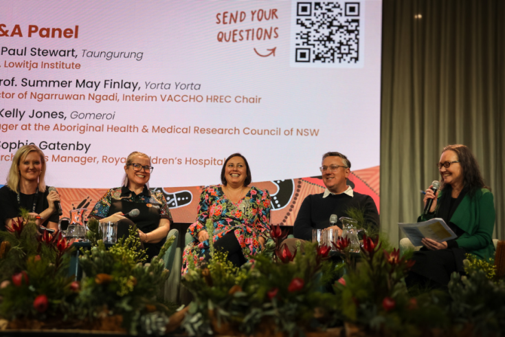 The image shows a panel of 5 individuals seated on stage with microphones. The individuals are Australian professionals in First Peoples health and research. Floral arrangements are visible in the foreground.