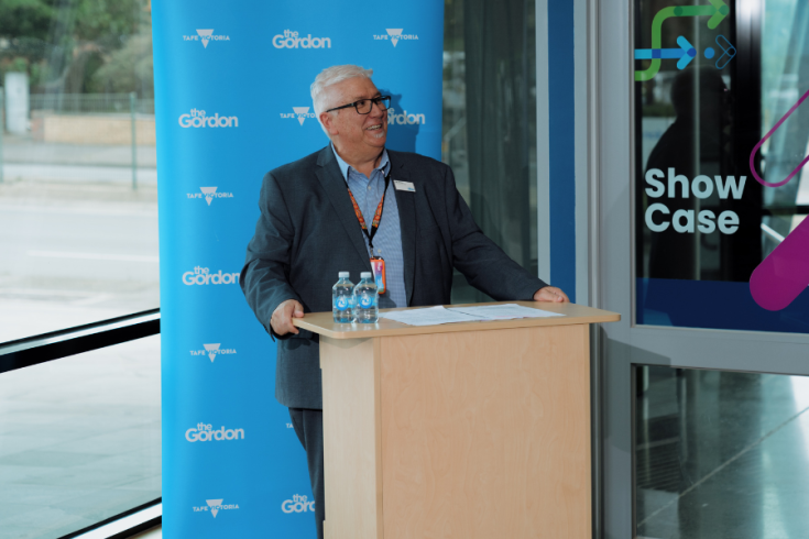 CEO of The Gordon Joe Ormeno stands at a podium with 2 water bottles and papers, speaking. They are in front of a blue backdrop featuring