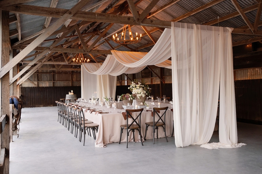 The interior of a rustic barn with a long dining table covered with a beige tablecloth and wooden chairs. Above the table, sheer white drapes hang from the rafters and chandeliers are on the roof. The table has floral centrepieces and place settings ready for an event.