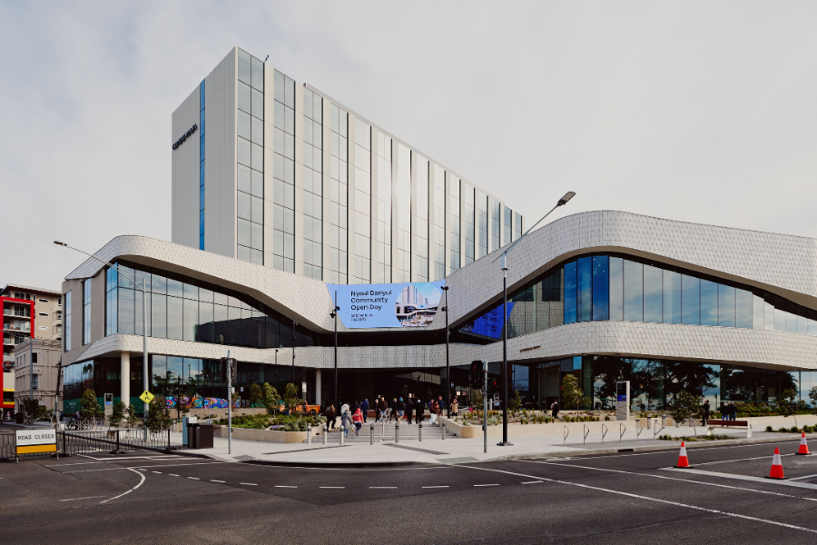 The modern, multi-story Nyaal Banyul building with a sleek, wavy design, featuring large glass windows and a digital screen. People are gathered near the entrance, with some entering the building. The area around has well-maintained landscaping and street infrastructure.