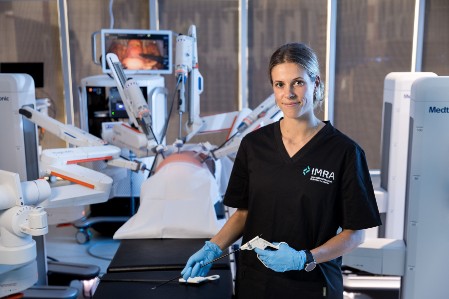 A person in an IMRA medical uniform holds medical equipment. In the background is an operating table with various pieces of medical equipment.