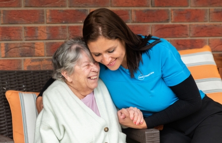 Diana Pasos in a blue uniform smiling at an elderly person in a white robe, both sitting on a bench.