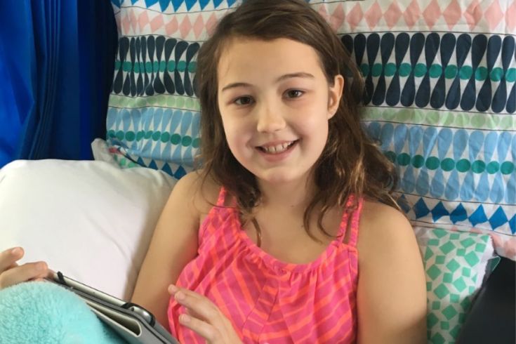 A person is smiling and looking at a tablet in a hospital bed with brightly coloured sheets. 