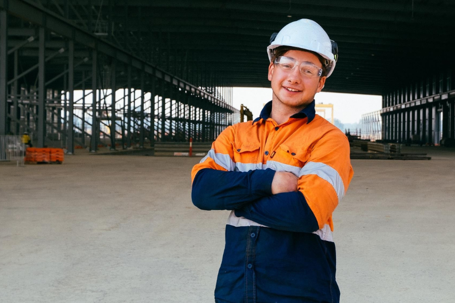 smiling construction worker wearing a hard hat and safety gear in front of an industrial setting