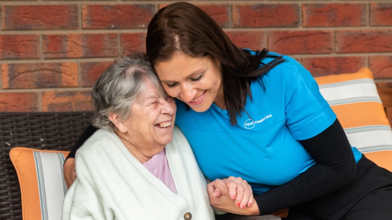 Diana Pasos in a blue uniform smiling at an elderly person in a white robe, both sitting on a bench.