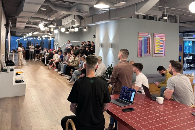 Attendees seated in rows at a tech talk event inside a modern office space, listening to a presenter near a digital screen.