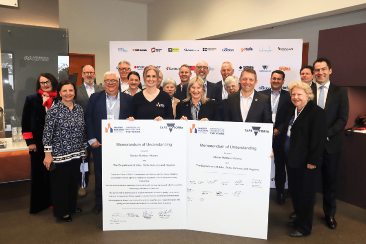 Group of individuals posing with a large signed document at the signing of a new agreement between Victorian Government and Master Builders Victoria with various company logos in the background.