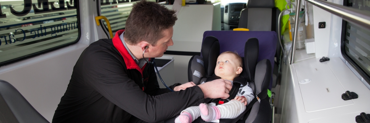 Staff member with stethoscope listening to chest of baby in baby seat in the back of the patient transport