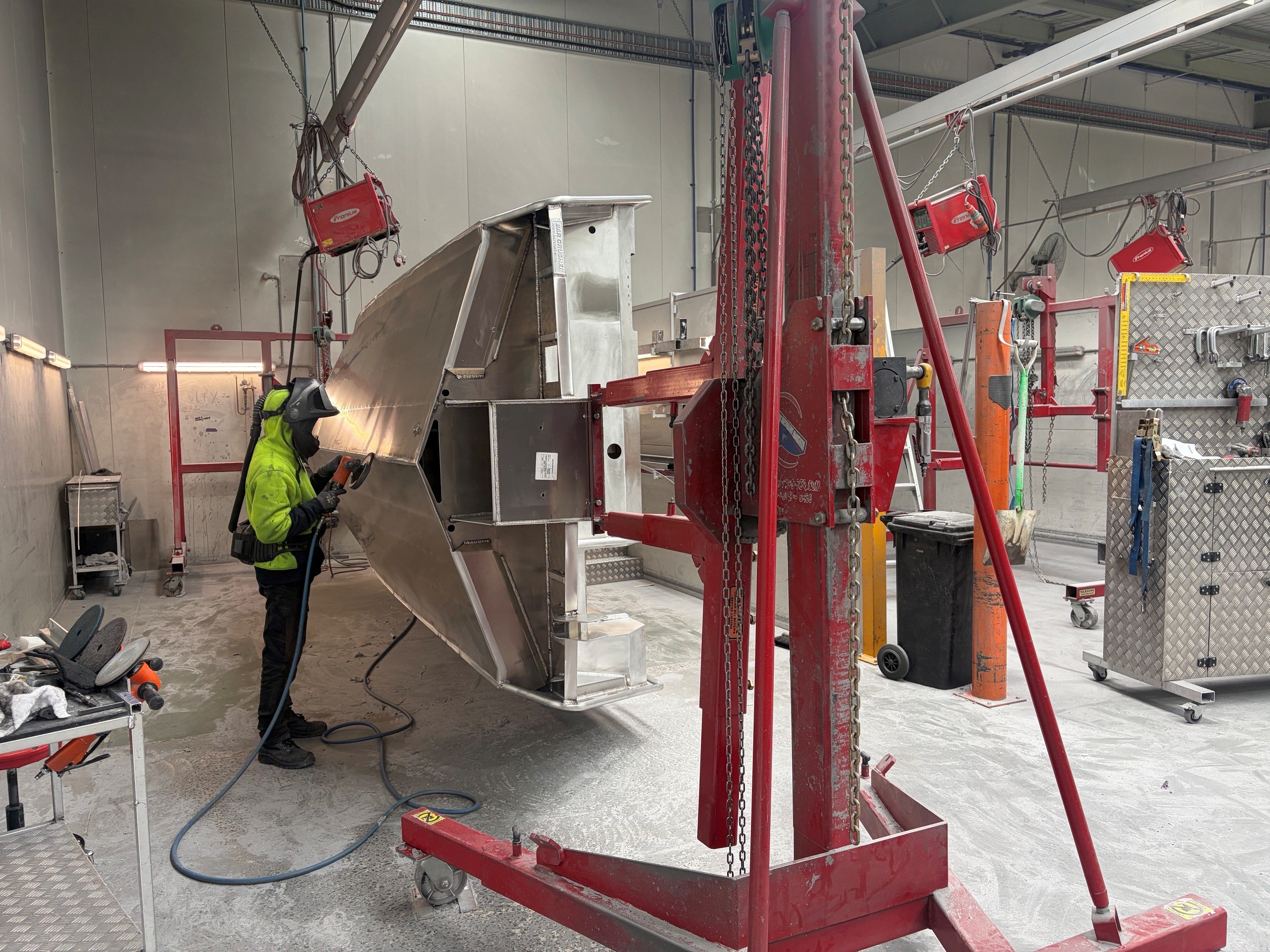 A person wearing a high visibility top is sanding a metal structure suspended by a red crane in a workshop that has industrial tools and machinery.
