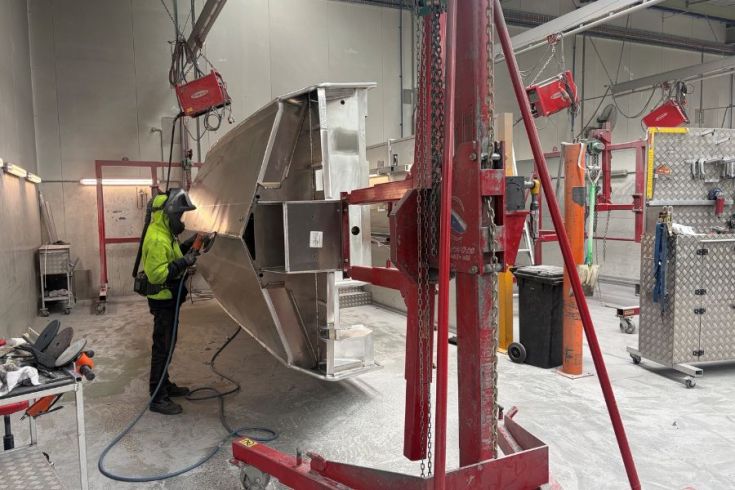 A person wearing a high visibility top is sanding a metal structure suspended by a red crane in a workshop that has industrial tools and machinery.