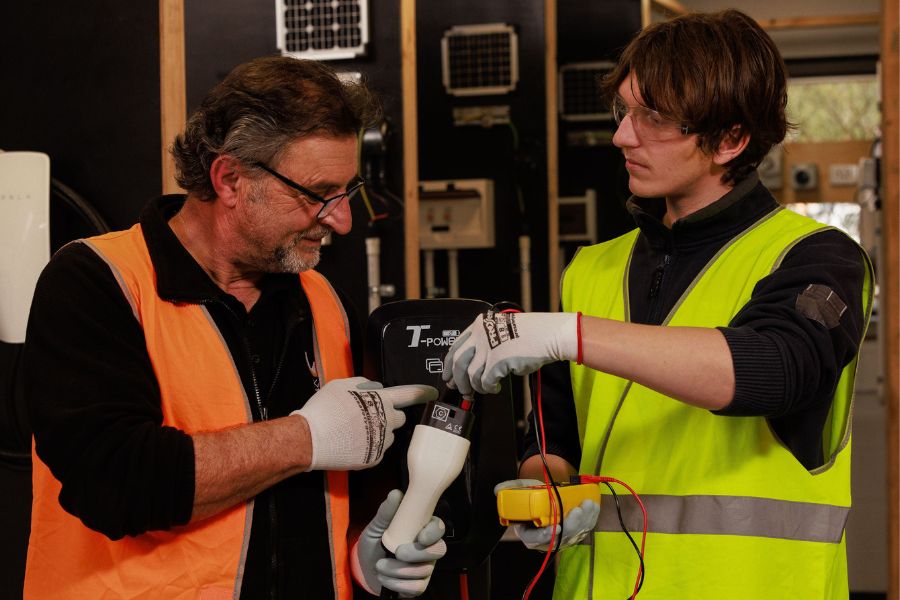 Two people are working together in a workshop. One is wearing an orange safety vest and gloves, pointing at a piece of equipment held by the other, who is in a yellow safety vest. Both are focused, with one holding a device and wires. They are surrounded by tools and machinery.
