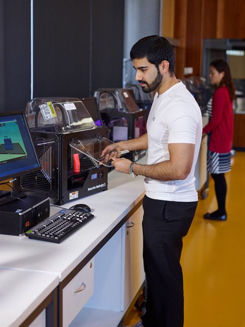 worker opens the door of a 3D printer to remove a red printed item while reviewing information on a computer screen