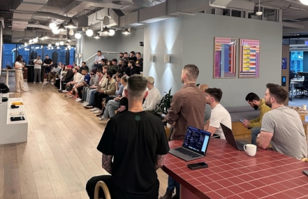 Attendees seated in rows at a tech talk event inside a modern office space, listening to a presenter near a digital screen.