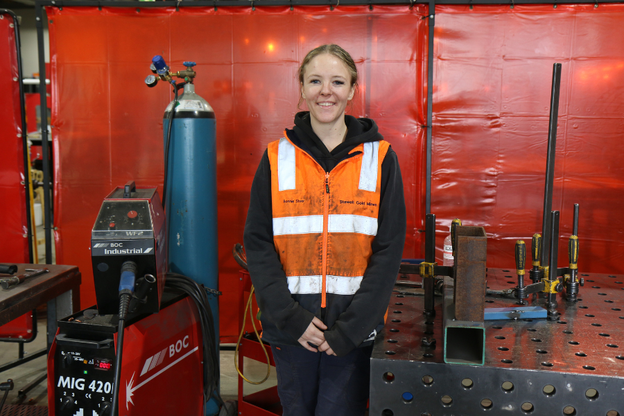 Bonnie Steer in protective gear proudly standing in front of welding equipment