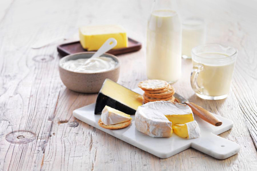 A table with various items on it: a selection of cheeses and crackers on a white cutting board, a bowl of yoghurt with a spoon, a cutting board with a slab of butter, and a bottle and jug of milk.