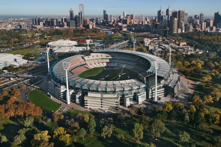 A view of the MCG stadium from above on a sunny day. The stadium has tiered seating and large floodlights. Melbourne can be seen in the background.