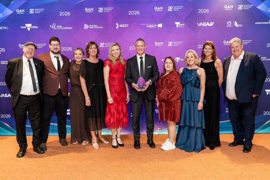 Ten people smile and pose in front of a purple media wall. One of them is holding an award. 