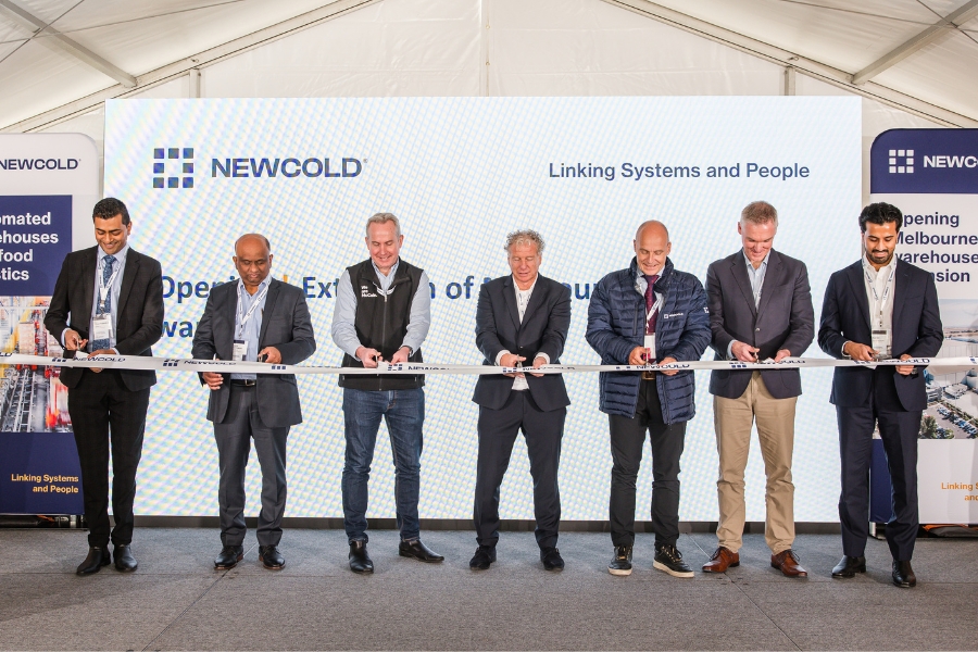 Seven people stand in front of a backdrop with NewCold logo, smiling and holding a white ribbon, suggesting a ribbon-cutting ceremony. 